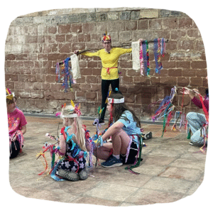 Colour photo of young people rehearsing inside an old priory building. They are dressed in bright colours with props like birds.