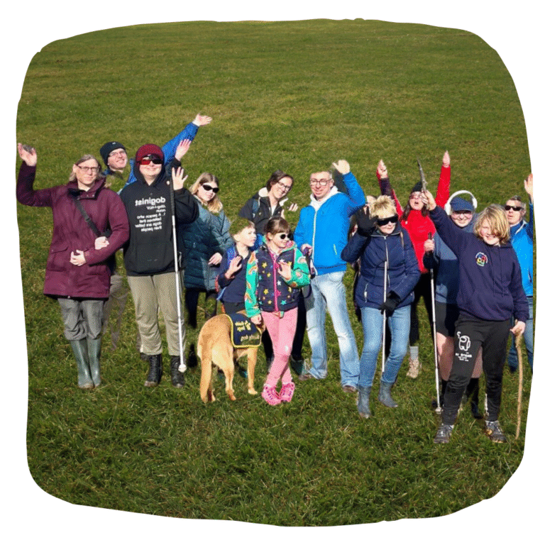 Colour image of people of all ages smiling and waving on a group walk, also featuring a buddy dog. Some people have canes. Behind them is a dark green grassy background.