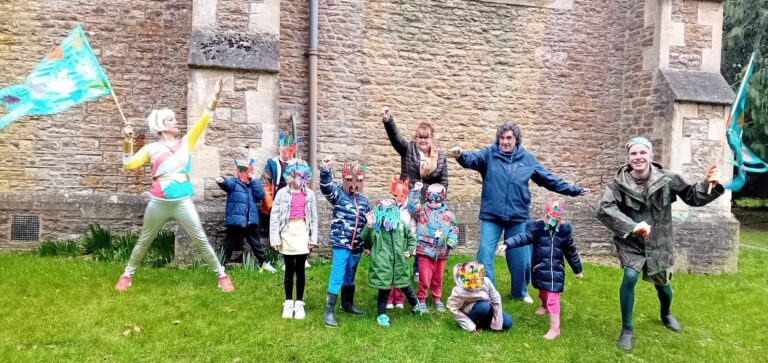 A group of adultS and children wearing masks and carrying flags. They are stood outside on grass, making Super Hero poses. A church building behind them.